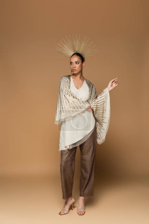 full length of brunette african american woman in shawl and golden crown posing on beige 