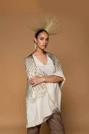brunette african american woman in shiny shawl and golden crown posing with crossed arms isolated on beige 