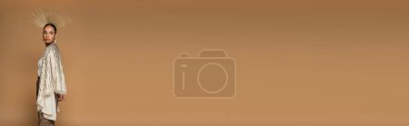 Téléchargez les photos : Brune afro-américaine en boucles d'oreilles et couronne dorée posant isolée sur beige, bannière - en image libre de droit