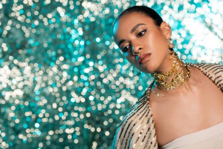 african american model with piercing and gold on neck posing while looking away on shiny blue background 