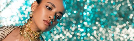 african american model with piercing and gold on neck posing while looking away on shiny blue background, banner
