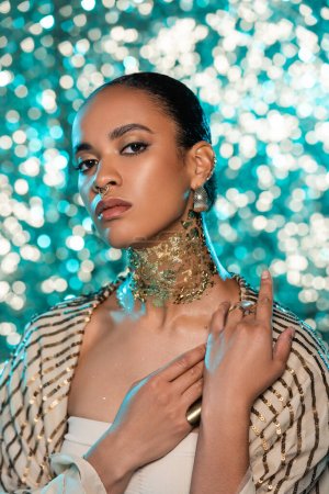 Afrikanische Amerikanerin mit Piercing und Gold am Hals posiert auf glitzerndem blauem Hintergrund 