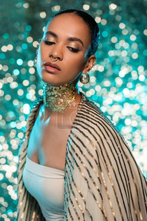pierced african american woman with jewelry and gold on neck posing on sparkling blue background 