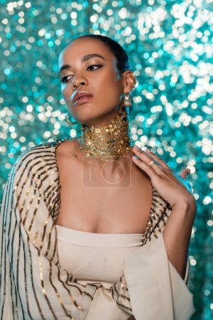 Téléchargez les photos : Femme américaine africaine sensuelle avec châle brillant et or sur le cou posant sur fond bleu étincelant - en image libre de droit
