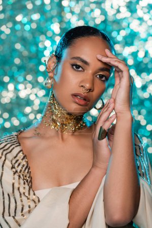 brunette african american woman in shawl and gold on neck looking at camera on sparkling blue background 