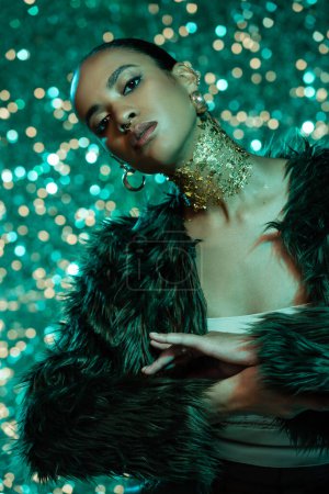 young african american woman in faux fur jacket and gold on neck posing on sparkling turquoise background 