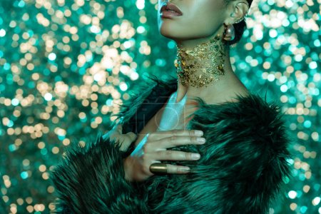 cropped view of african american woman in faux fur jacket and gold on neck posing with crossed arms on turquoise background 