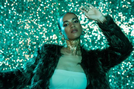 pretty african american woman in faux fur jacket and gold on neck posing on shiny blue background 