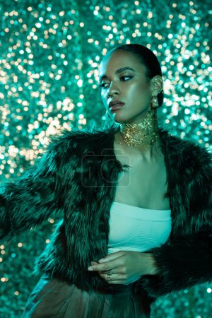 stylisches afrikanisch-amerikanisches Model in Kunstpelzjacke und Gold am Hals posiert auf blauem Hintergrund 