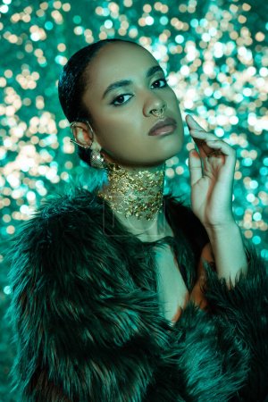 brunette african american woman in trendy faux fur jacket and gold on neck looking at camera on shiny blue background 