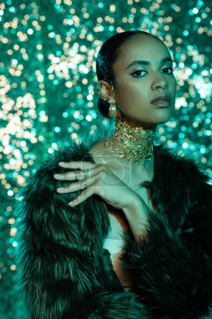 Photo for Stylish african american woman in faux fur jacket and golden foil on neck looking at camera on sparkling background - Royalty Free Image