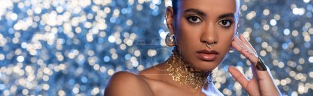 Trendy african american model in golden accessories on sparkling blue background, banner 