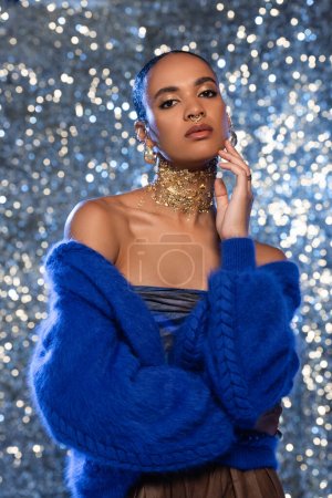 Stylish african american model in sweater and accessories touching face on sparkling background 
