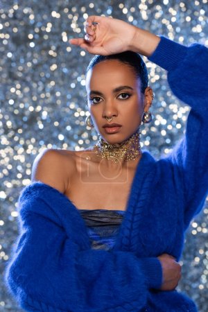 Trendiges afrikanisch-amerikanisches Model in blauem Pullover und goldenen Accessoires auf glitzerndem Hintergrund 