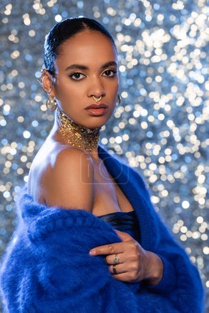 Stilvolle afrikanisch-amerikanische Frau mit goldenen Accessoires trägt blauen Pullover auf glitzerndem Hintergrund 