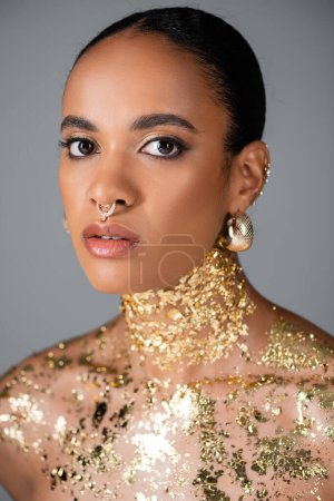 Mujer afroamericana de moda con accesorios dorados y lámina en el pecho aislada en gris 