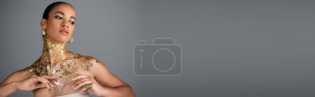 Foto de Joven modelo afroamericano con lámina dorada en el pecho mirando hacia otro lado aislado en gris, pancarta - Imagen libre de derechos