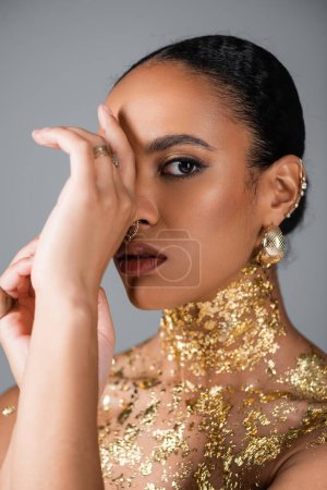 Téléchargez les photos : Femme afro-américaine avec maquillage doré et feuille sur la poitrine posant isolée sur gris - en image libre de droit