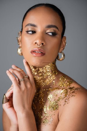 Young african american woman with golden makeup and foil on chest looking away isolated on grey 