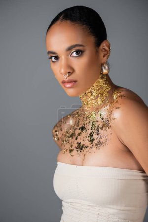 Téléchargez les photos : Femme afro-américaine à la mode avec percing et feuille d'or sur la poitrine en regardant la caméra isolée sur gris - en image libre de droit