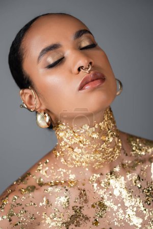 Téléchargez les photos : Modèle afro-américain avec accessoires dorés et feuille sur les yeux de fermeture du corps isolés sur gris - en image libre de droit