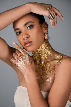 Téléchargez les photos : Femme afro-américaine à la mode avec accessoires et feuille d'or sur le corps et les mains posant isolé sur gris - en image libre de droit