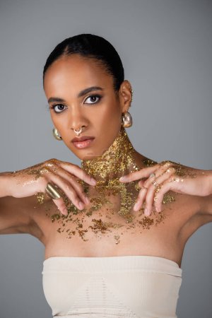 Téléchargez les photos : Femme afro-américaine à la mode avec feuille sur le corps et accessoires dorés posant isolé sur gris - en image libre de droit