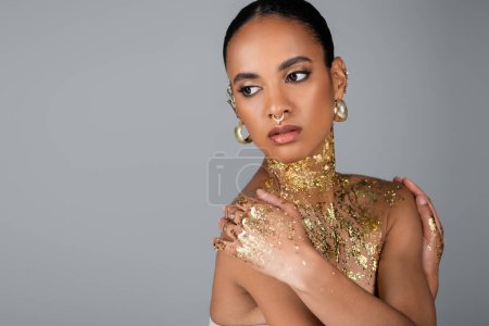 Téléchargez les photos : Femme afro-américaine élégante avec feuille d'or sur le corps touchant les épaules isolées sur gris - en image libre de droit