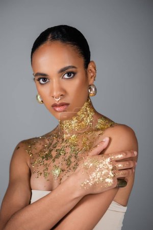 Modisches afrikanisch-amerikanisches Model mit goldener Folie auf der Brust und Piercing, das isoliert auf grau in die Kamera blickt 