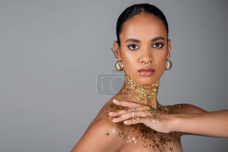 Trendy afrikanisch-amerikanische Frau mit goldener Folie an Hals und Brust posiert isoliert auf grau 