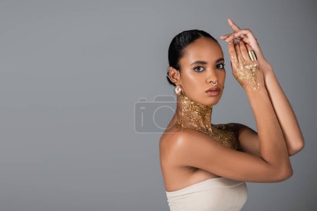 Stylish african american woman in accessories and golden foil on body looking at camera isolated on grey 