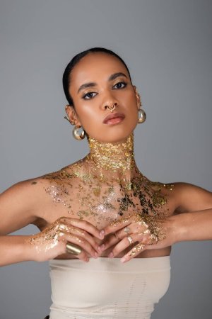 Téléchargez les photos : Femme afro-américaine à la mode avec feuille d'or sur le corps et les mains posant isolé sur gris - en image libre de droit