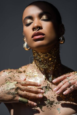 Trendy african american woman with golden foil on body standing in light isolated on grey 