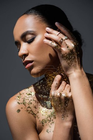 Téléchargez les photos : Jeune femme afro-américaine avec feuille d'or sur le corps touchant le visage tout en posant dans la lumière isolée sur le gris - en image libre de droit