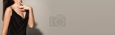 Cropped view of brunette woman in black dress touching neck on grey background, banner 