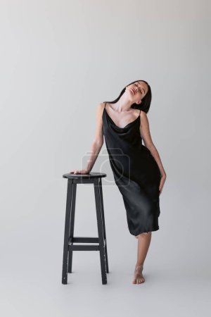 Sinnliche Frau mit Vitiligo im Seidenkleid berührt Stuhl auf grauem Hintergrund 