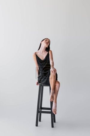 Zufriedene Frau mit Vitiligo überkreuzten Beinen, während sie auf Stuhl auf grauem Hintergrund sitzt 