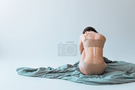 vista posterior de la mujer morena con condición de piel crónica vitiligo sentado en manta sobre fondo gris 