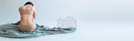 back view of brunette woman with vitiligo chronical skin condition sitting on blanket on grey background, banner