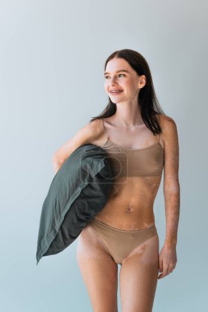 happy young woman with vitiligo and braces standing in beige lingerie and holding pillow isolated on grey