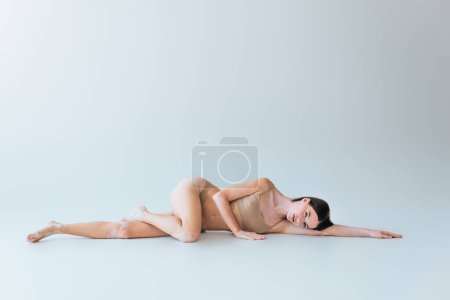 Photo for Full length of barefoot and young woman with vitiligo lying in beige lingerie on grey background - Royalty Free Image