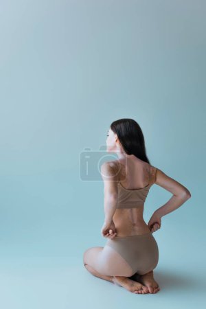 full length of barefoot and young woman with vitiligo sitting and adjusting beige panties on grey