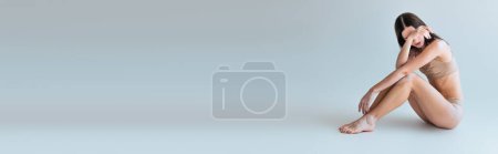 Téléchargez les photos : Pleine longueur de pieds nus et jeune femme avec vitiligo assis en lingerie beige et couvrant le visage sur gris, bannière - en image libre de droit