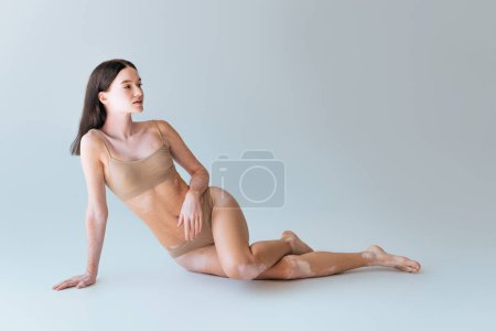 full length of brunette young woman with vitiligo sitting in beige lingerie on grey