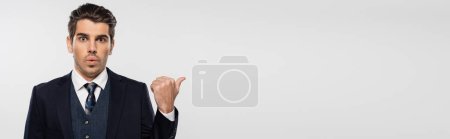 surprised businessman in suit pointing with thumb away isolated on grey, banner
