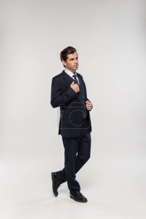Foto de Longitud completa de buen aspecto y joven hombre de negocios en traje ajustando chaqueta en gris - Imagen libre de derechos