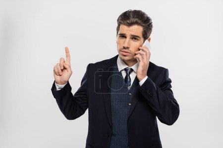 joven hombre de negocios en traje apuntando hacia arriba y hablando en el teléfono inteligente aislado en gris