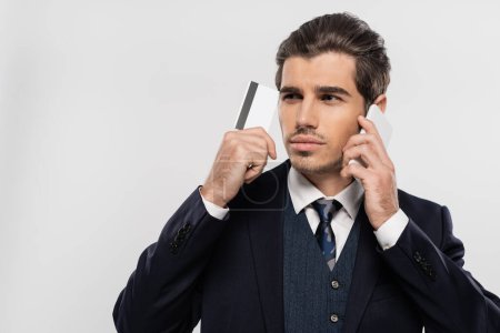 young businessman in suit talking on smartphone and holding credit card isolated on grey 