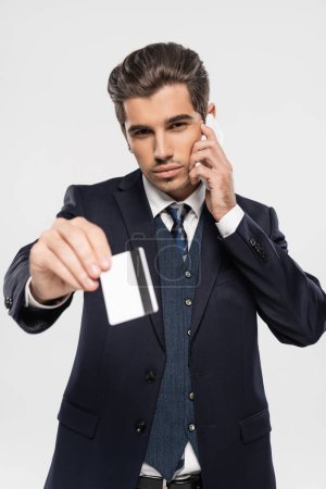 Selbstbewusster Geschäftsmann im Anzug spricht auf Smartphone und hält Kreditkarte isoliert auf grau 
