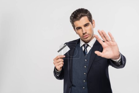 erfolgreicher Geschäftsmann im Anzug hält Kreditkarte und zeigt Stopp-Geste isoliert auf grau